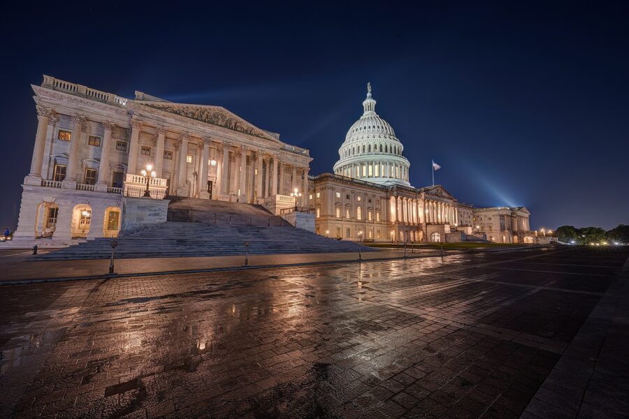 The United States Capitol at night, its dome and colonnaded wings lit against a dark sky and reflected in the wet stone plaza.